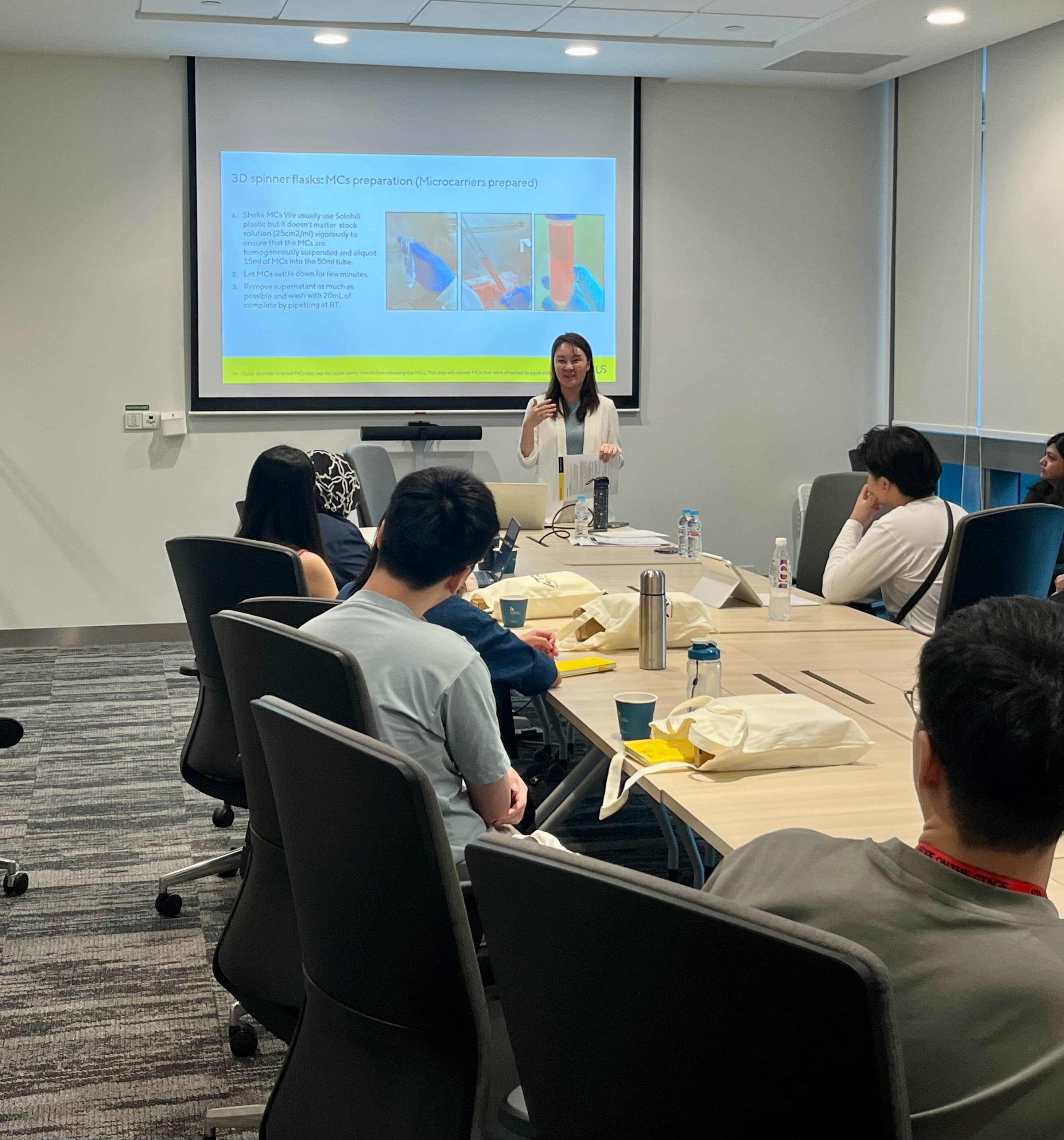 Facilitator delivering a presentation on microcarrier preparation for 3D spinner flasks during an ACTRIS-Sartorius workshop, with participants seated in a conference room attending the training session.
